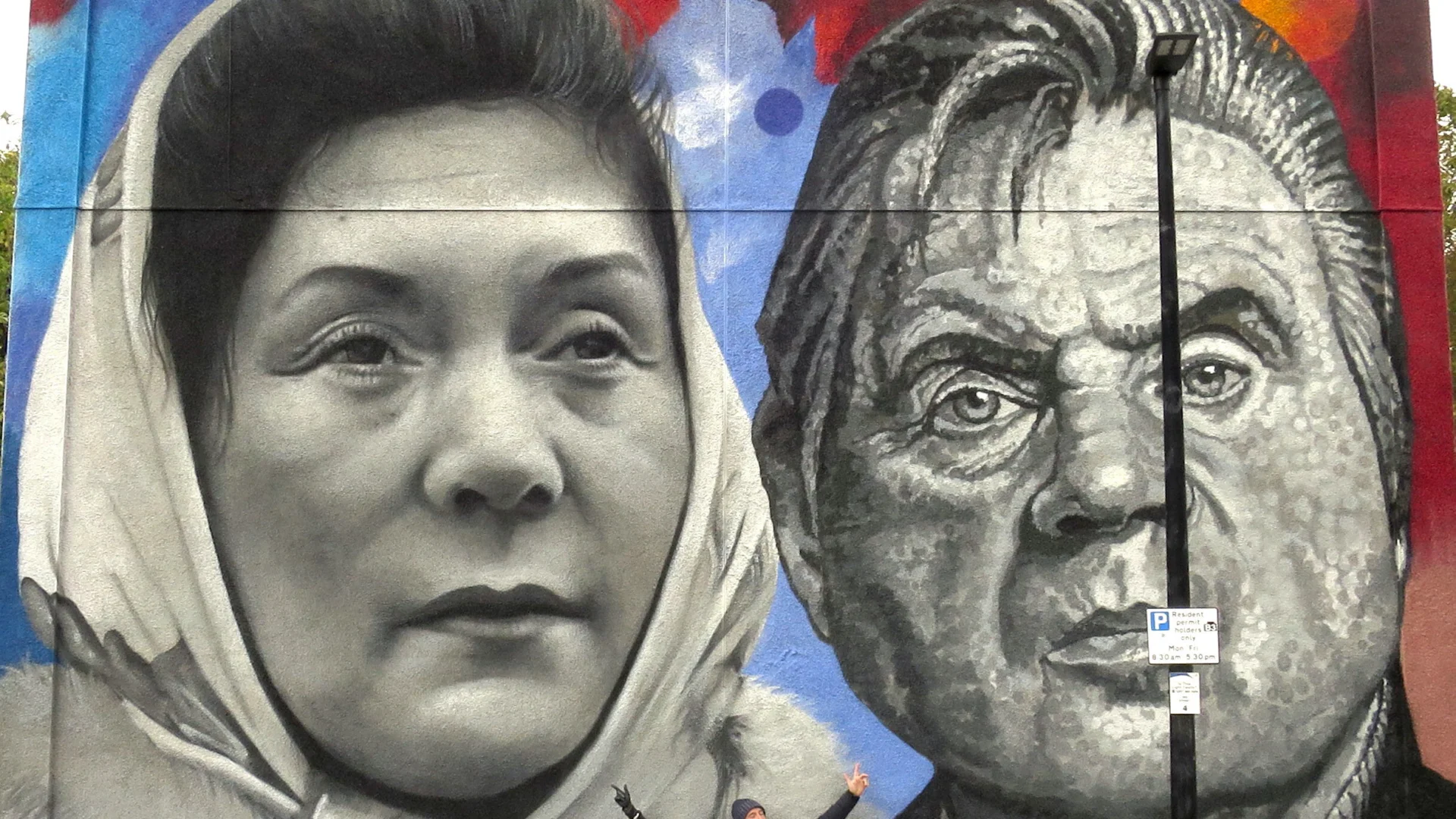 Two people standing in front of a large mural of two faces.