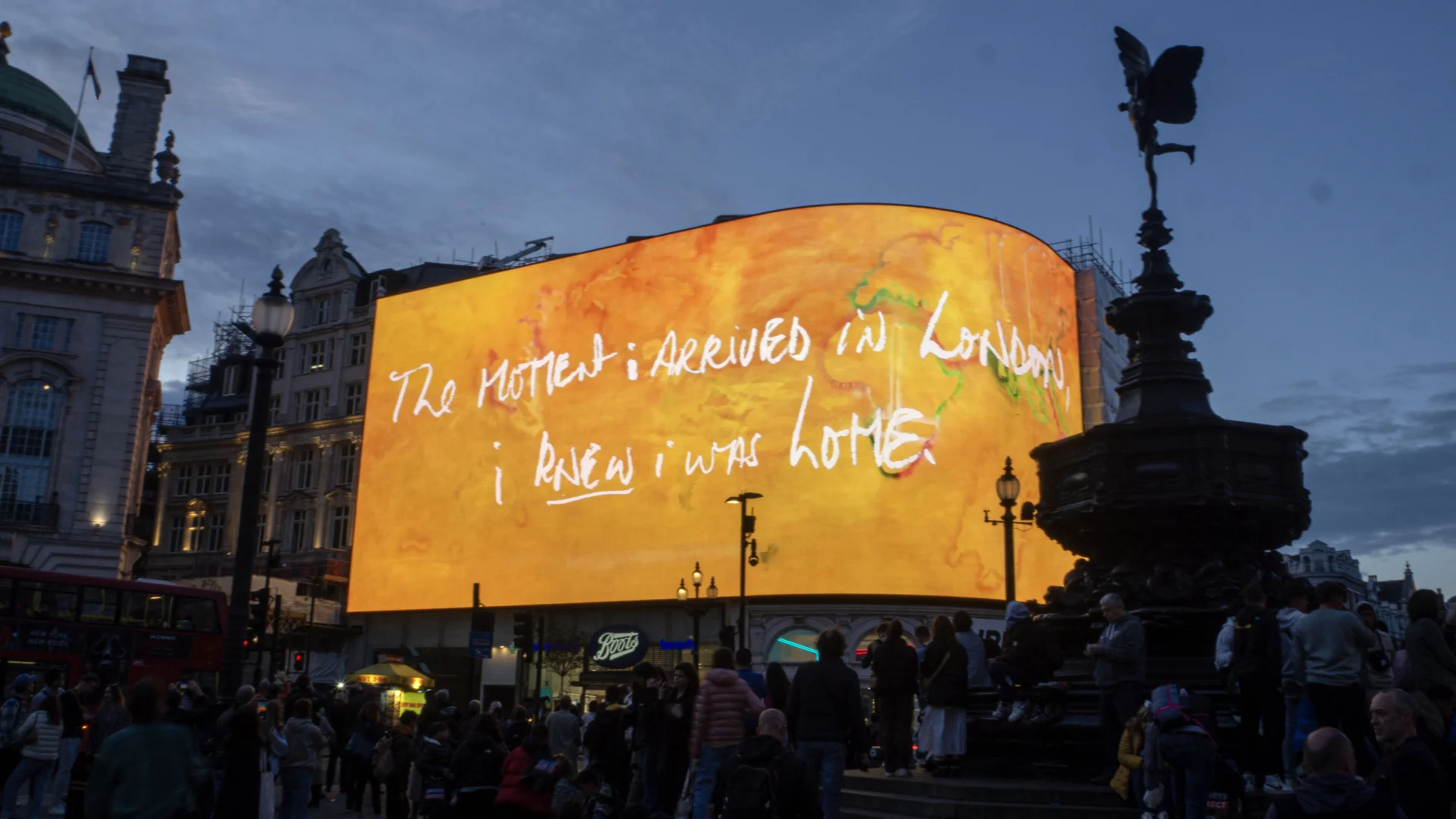The billboard in Piccadilly Circus, London, showing an abstract artwork in orange hues, overlaid with the text 'The moment I arrived in London, I knew I was home