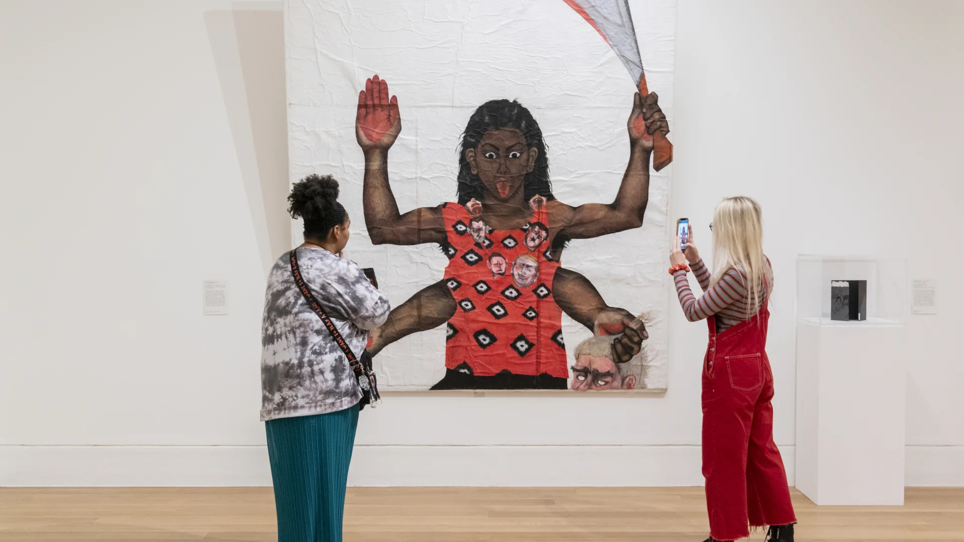 Two people in front of an artwork of a brown woman on a red shirt with four arms, one hand hidden behind the viewer, one hand holding a knife, one hand holding a head and one with blood on it.