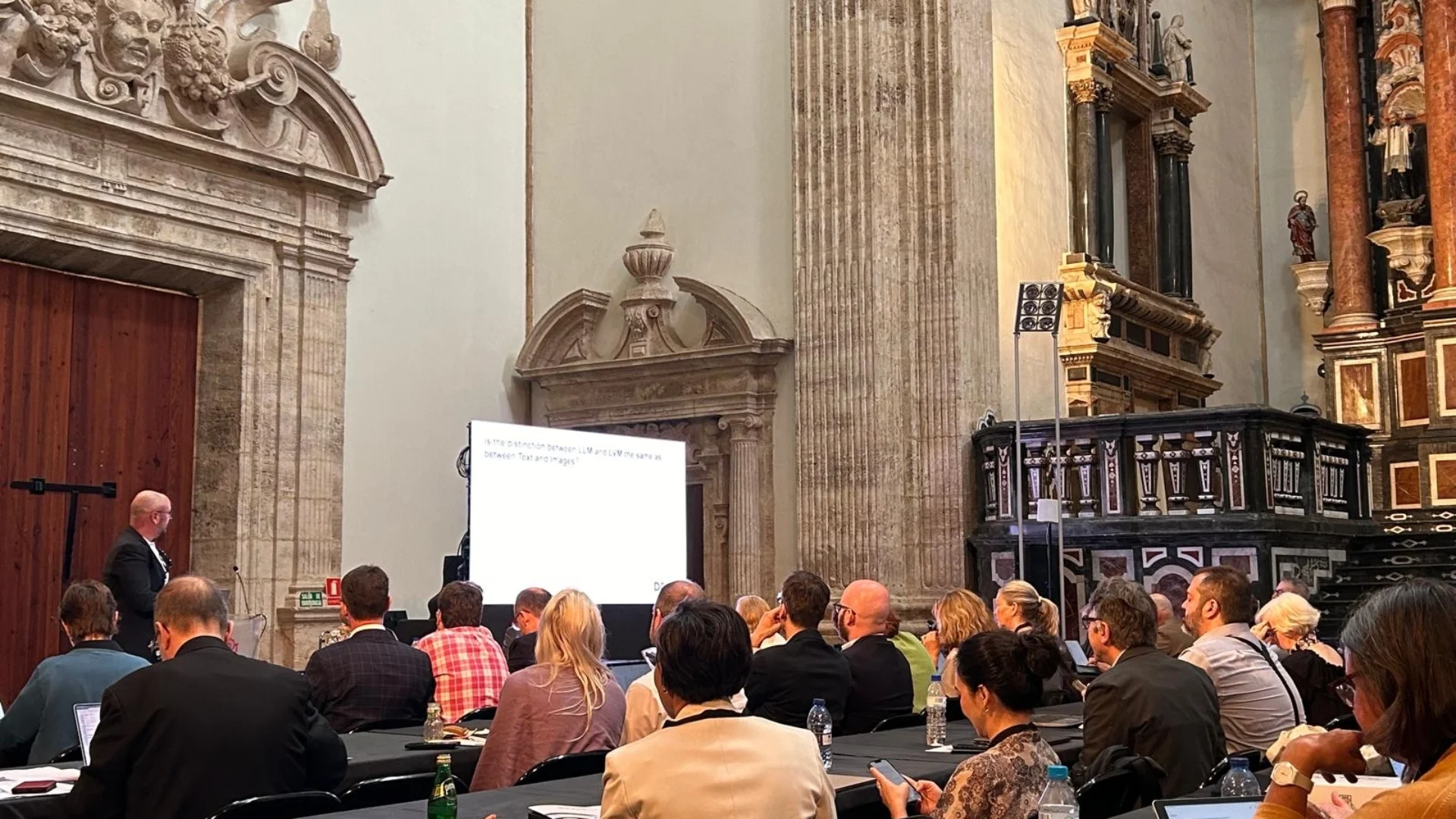 People sitting in front of a screen projection of a presentation in a cathedral-like room.