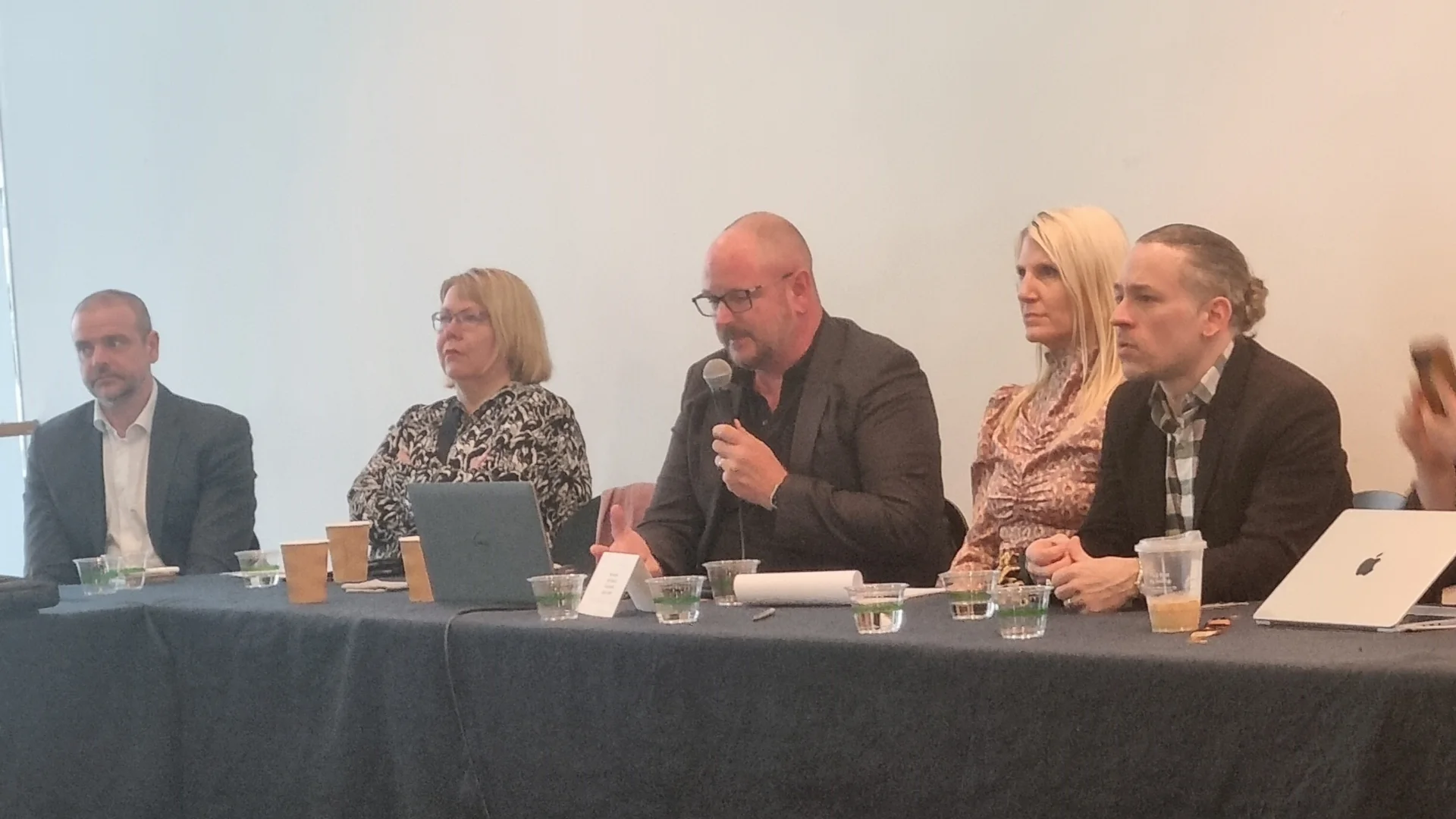 A panel of people, sitting at a table, with the people in the middle holding a microphone and speaking