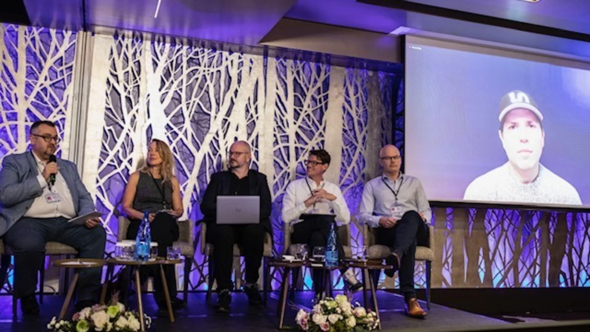 Four people sitting on a stage in front of a screen of someone video-calling them