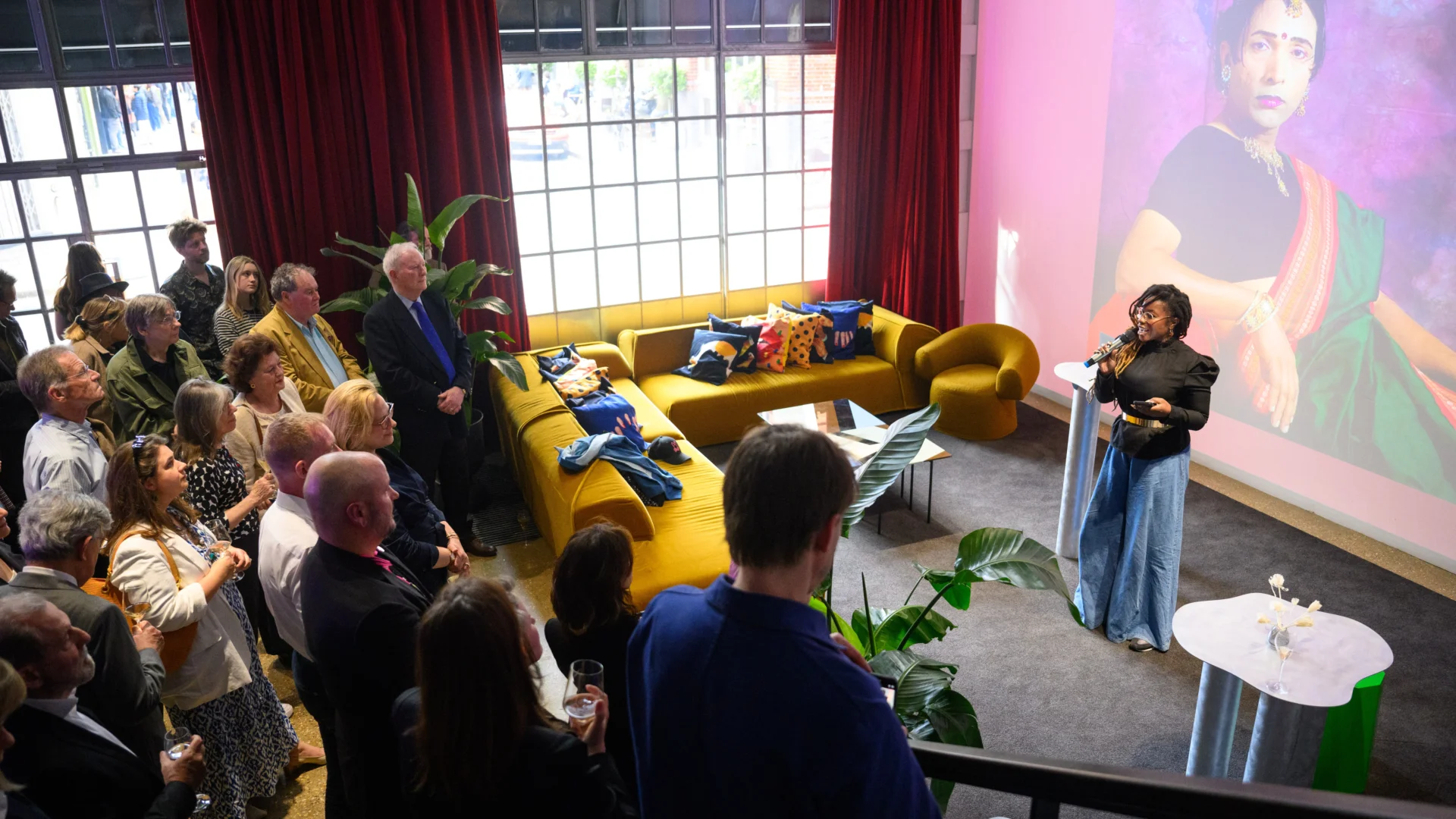 A room full of people, circling a person who is giving a speech in front of a big screen