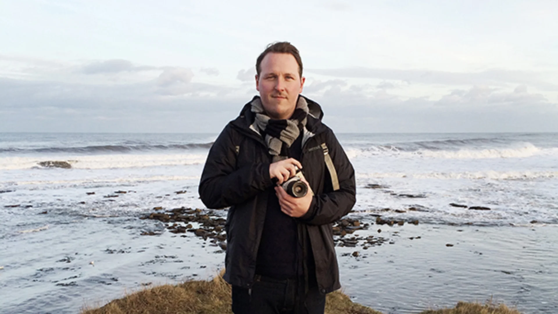 Owen Richards standing holding a camera in front of waves breaking on a rocky beach
