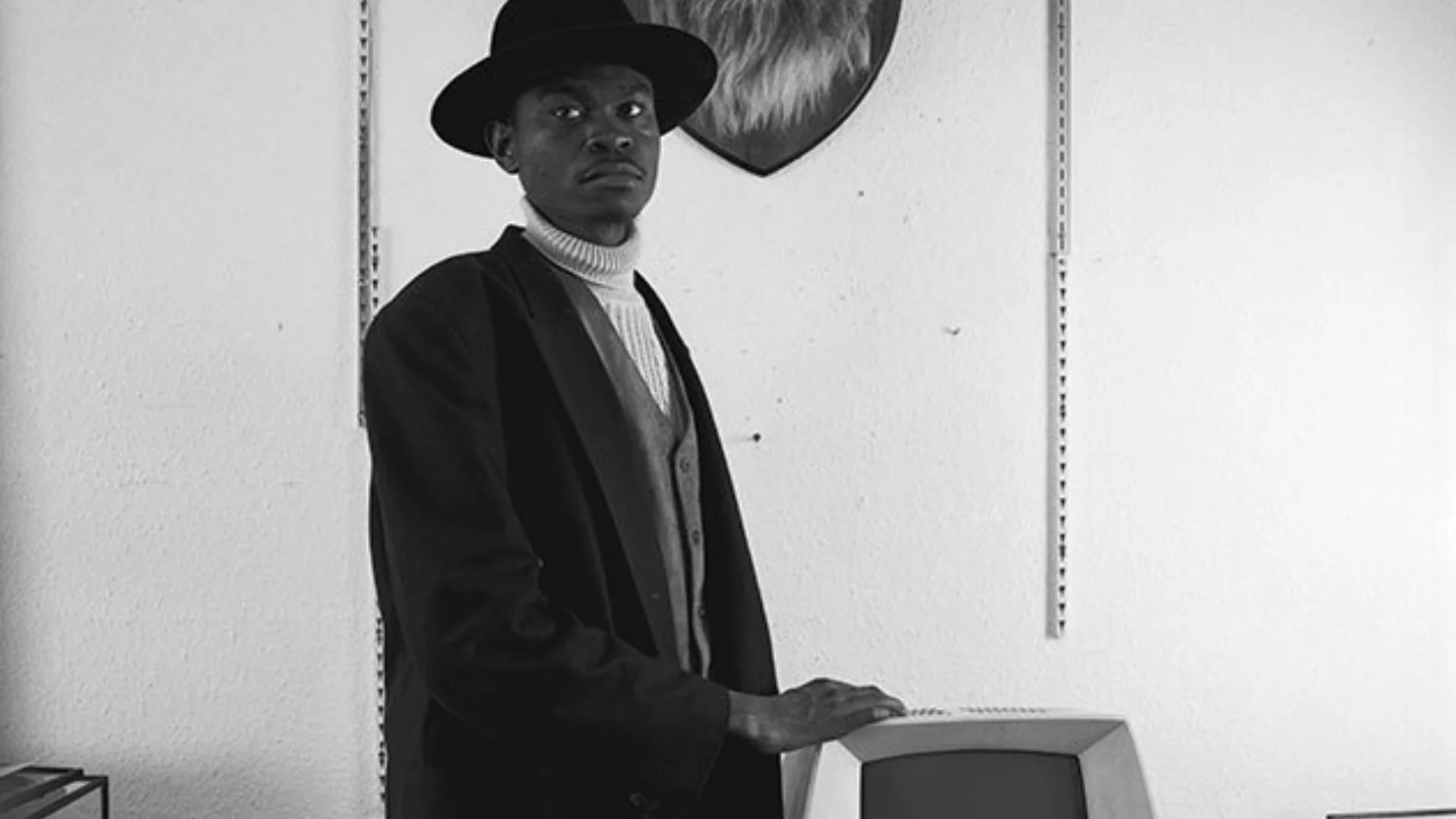 A man in a hat standing next to an old PC and in front of a stuffed and wall mounted lions head