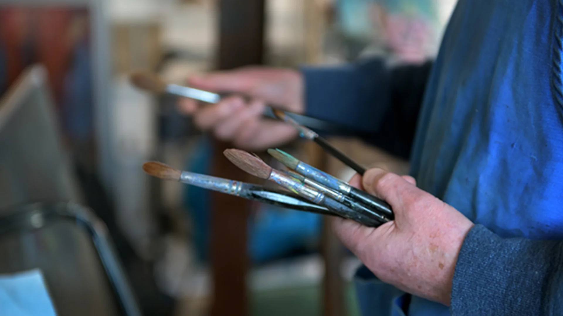 Painters hands pictured holding paint brushes facing a canvas