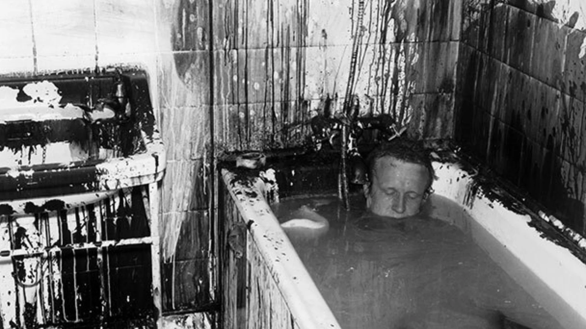 Black and white photo of man submerged in bath tub. Walls of bathroom covered in a dark paint