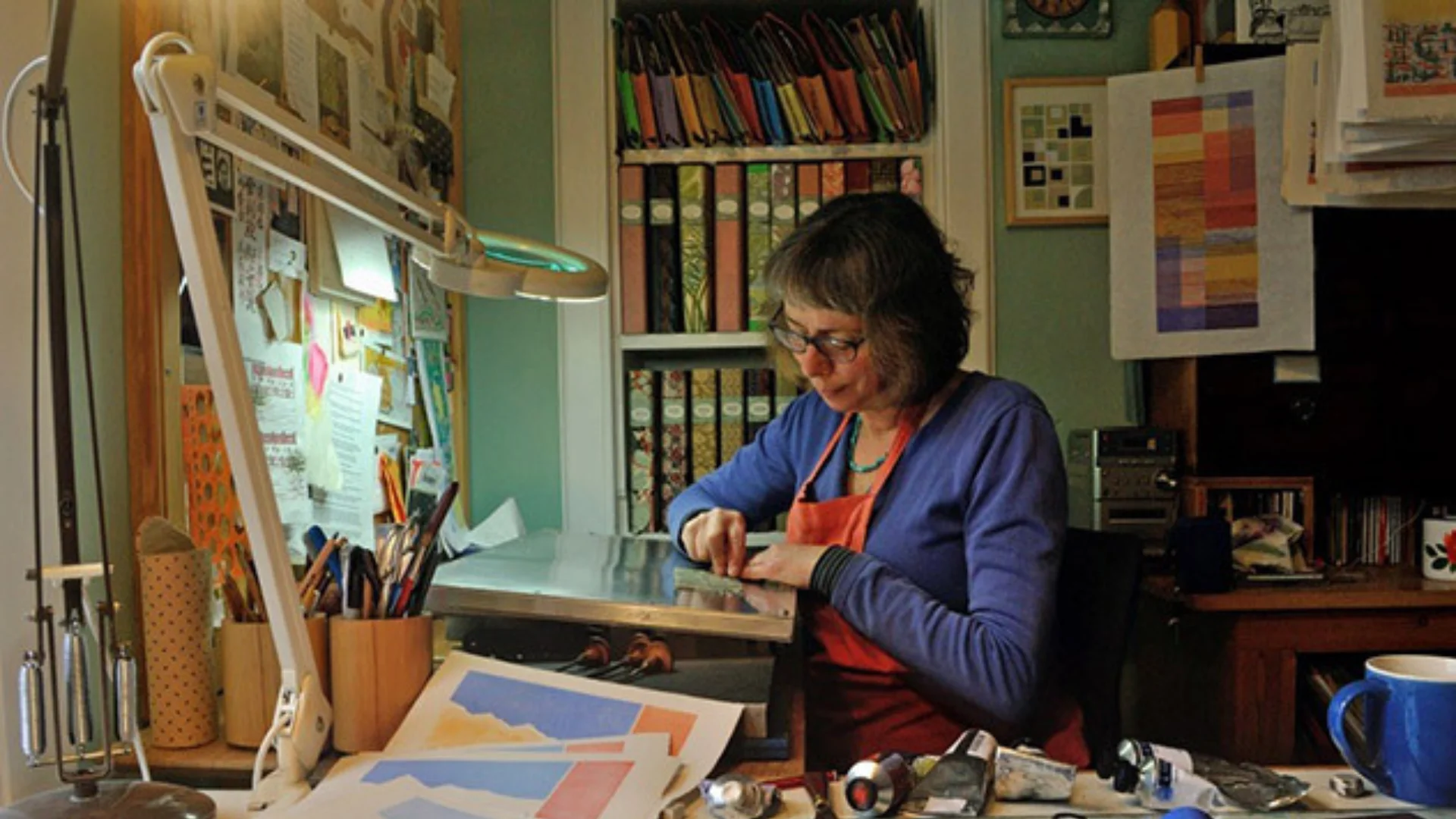 A white woman sitting on a desk, surrounded by paint and artworks