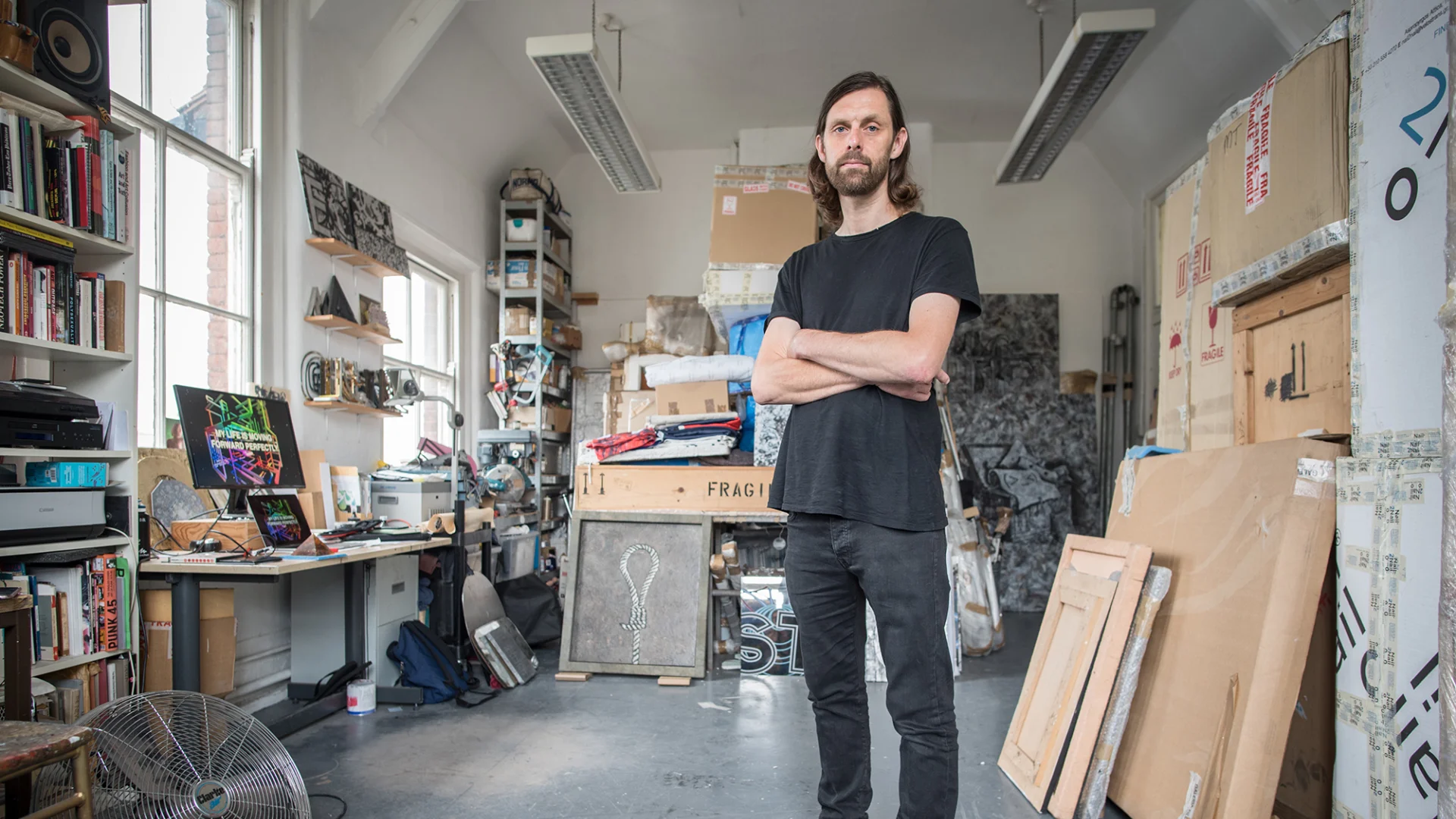 A white man with brown long hair and a beard standing in the middle of his art studio