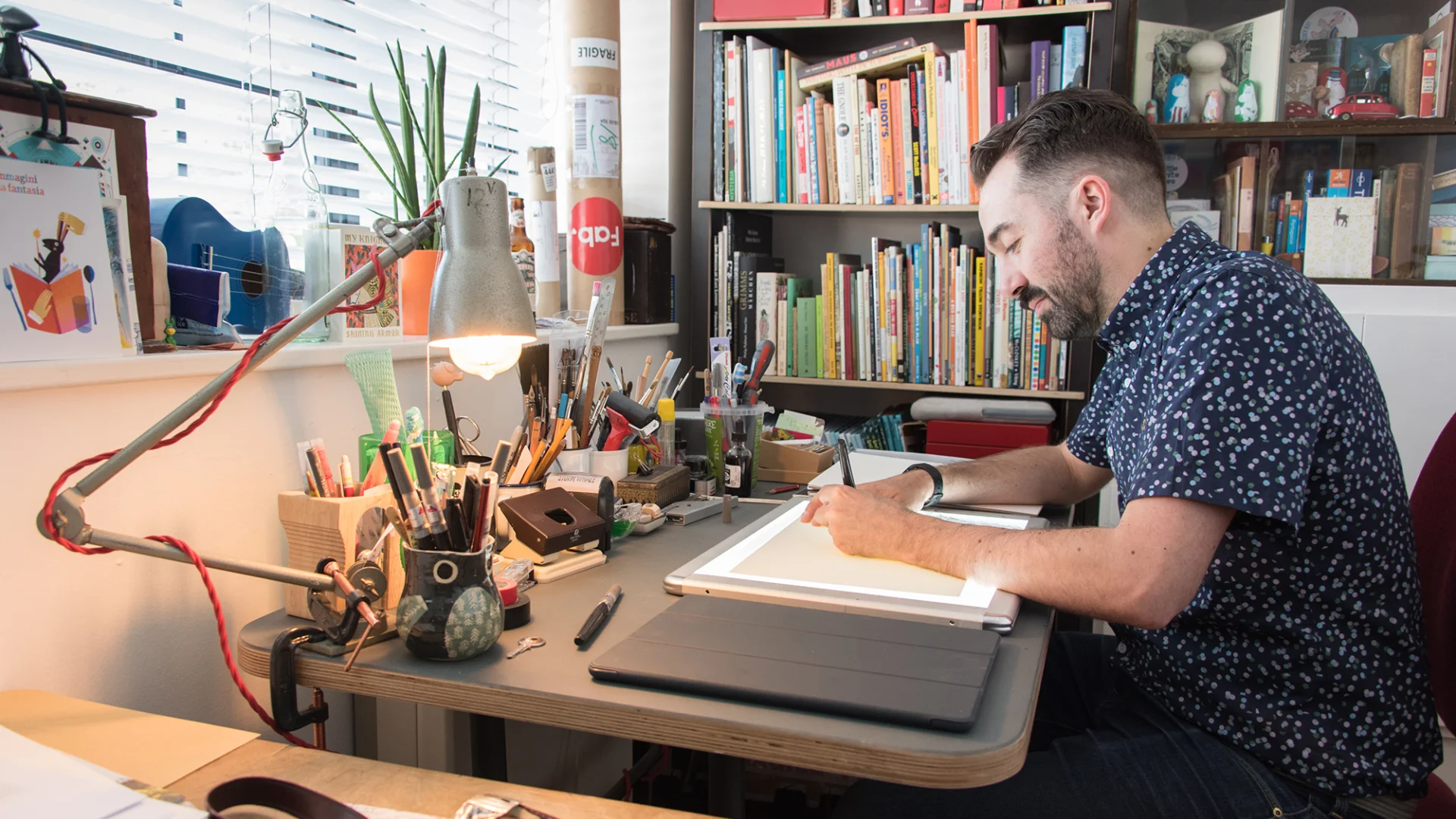 A white man sitting on a desk and drawing next to a book case
