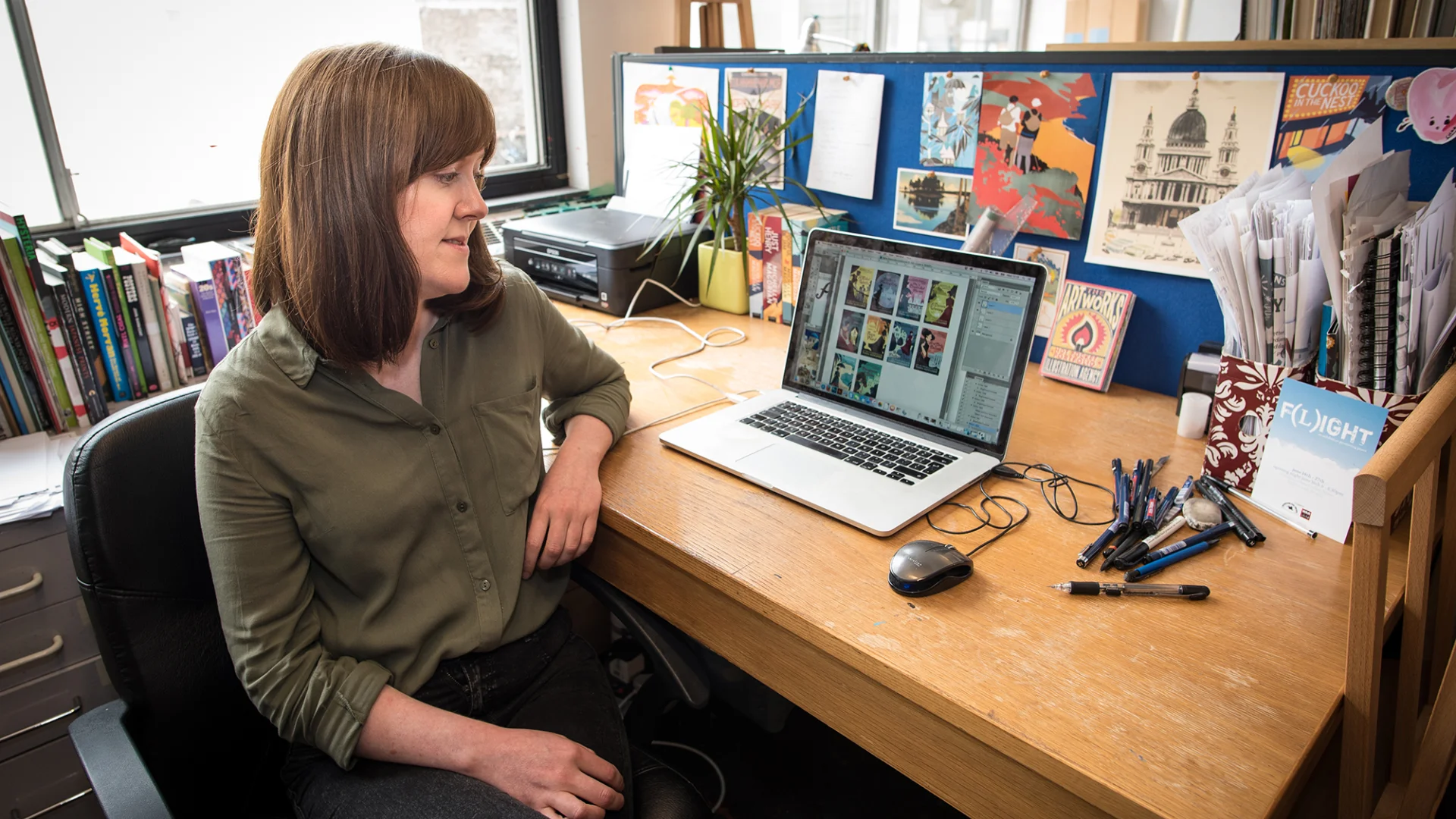 A white woman sitting, surrounded by her artworks on a desk, looking at a computer