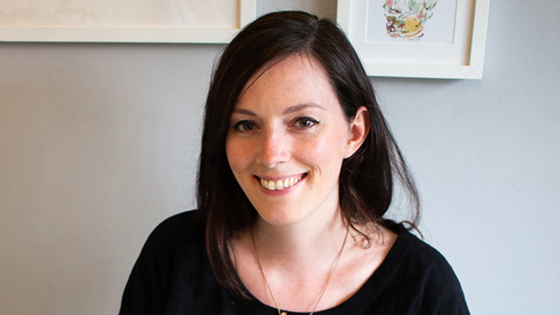 A white woman with black hear sitting on a desk, looking at the camera