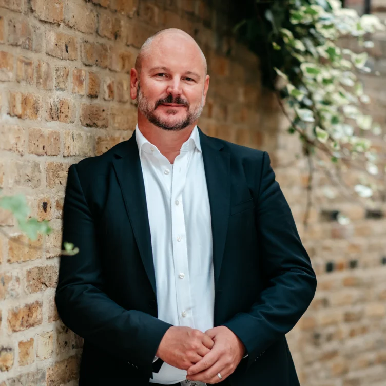 A white man in a suit standing in front of a brick wall