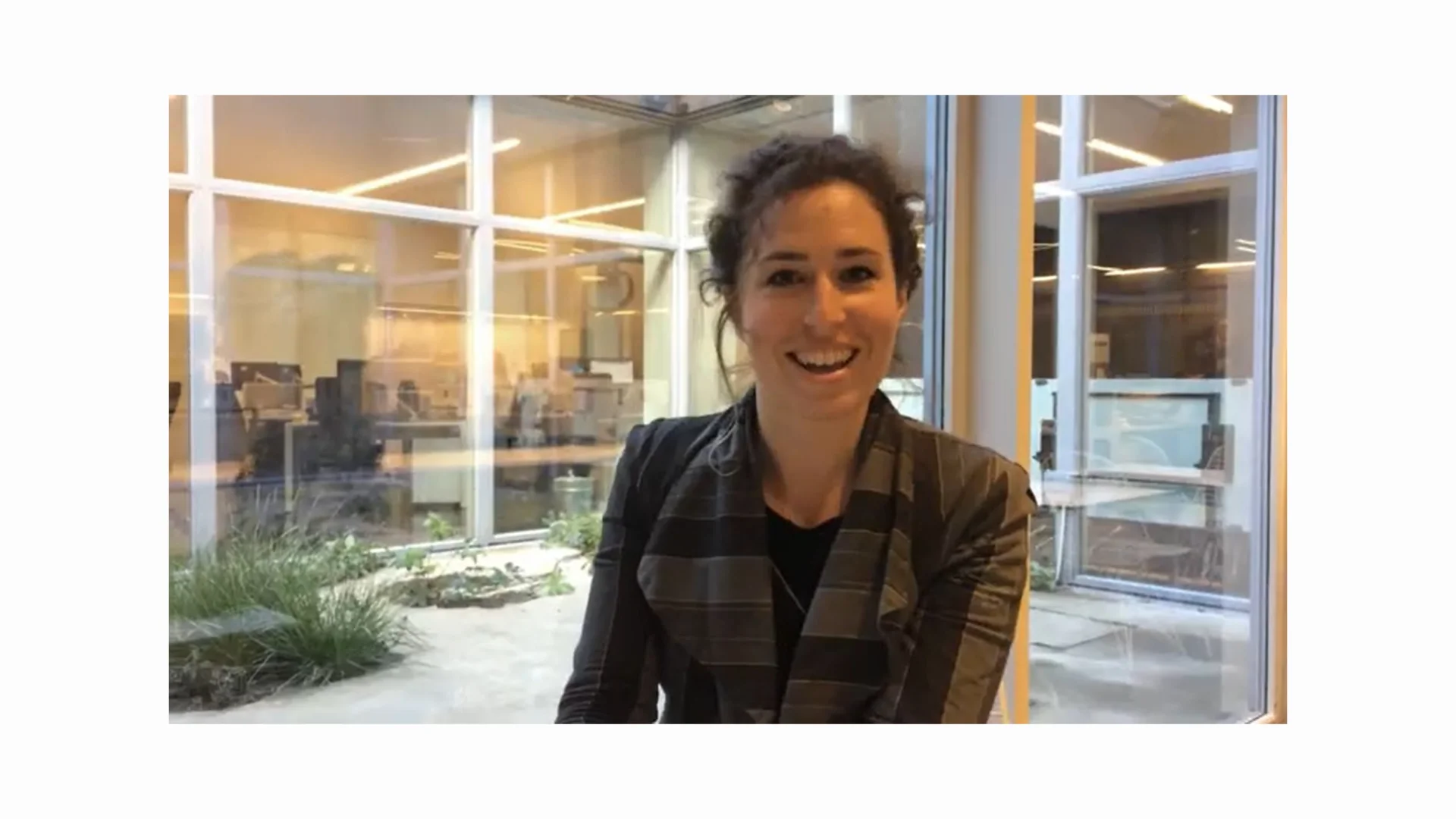 A white woman with brown hair in a bun sitting in an office and speaking at the camera