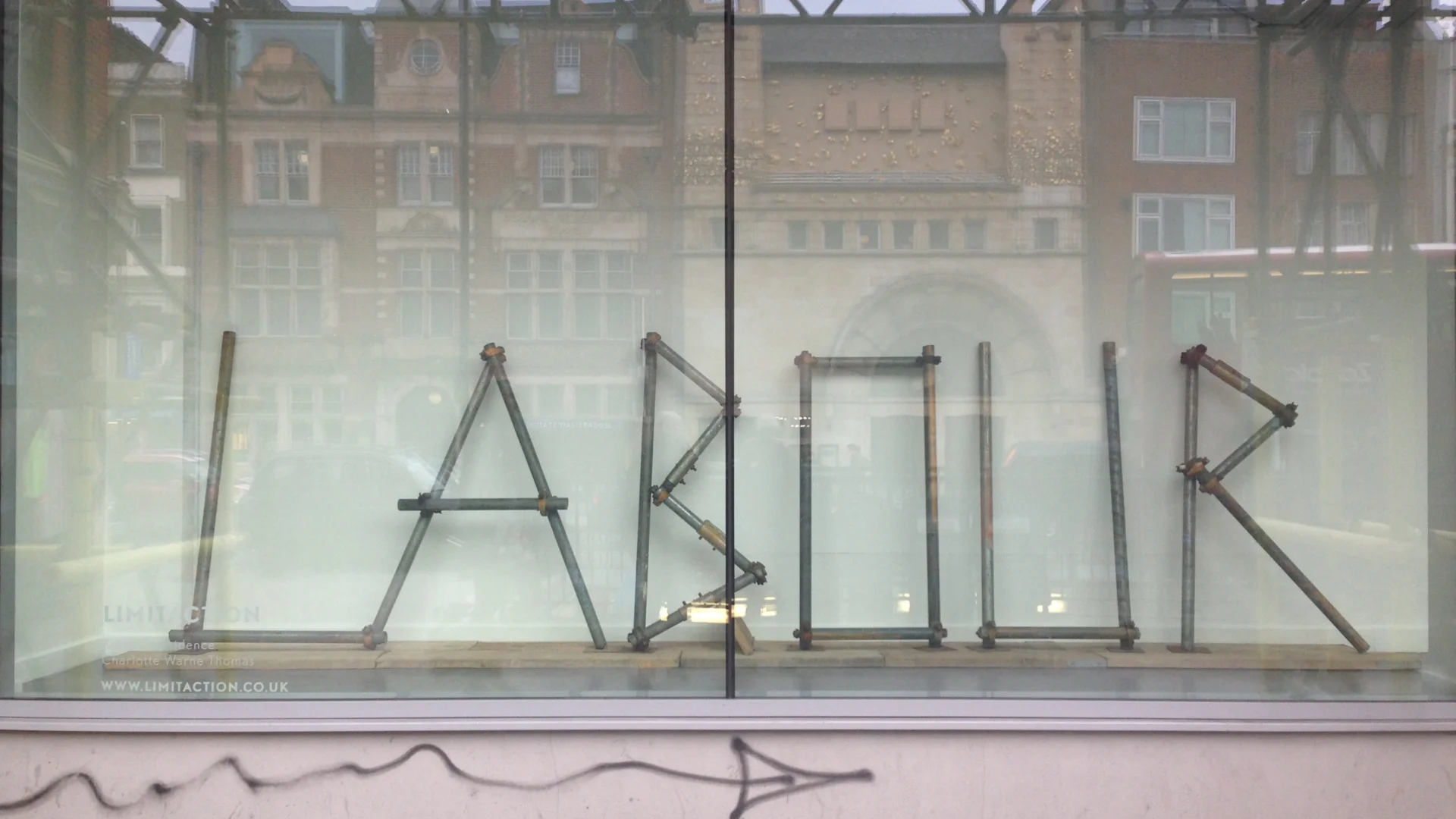 A photograph of an artwork installation. The work spells our the word LABOUR, using scaffolding poles.