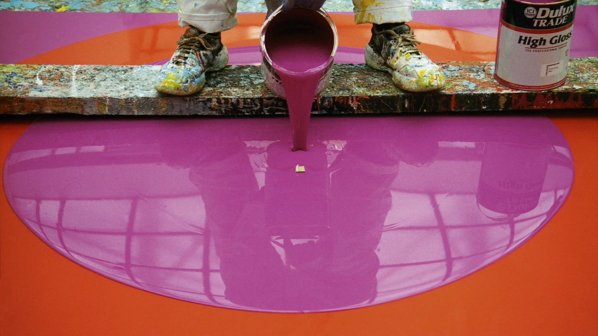 A colour photograph showing magenta paint being poured from a paint can onto an orange canvas.